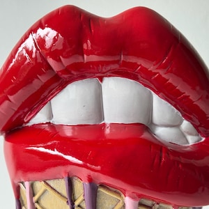 Könnte beinhalten: Eine glänzende, rote Skulptur offener Lippen mit weißen Zähnen. Die Lippen sind mit einem glänzenden Finish versehen, und die Basis weist ein Waffelkonus-Design mit tropfenden rosa und lila Akzenten auf. Ein einzigartiges Dekorationsstück.