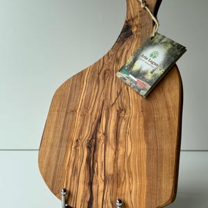 May include: A wooden cutting board with a handle, featuring a natural wood grain pattern. The board has a tag that reads "Arte Legno" and "Product of Italy". The board is supported by two metal stands.
