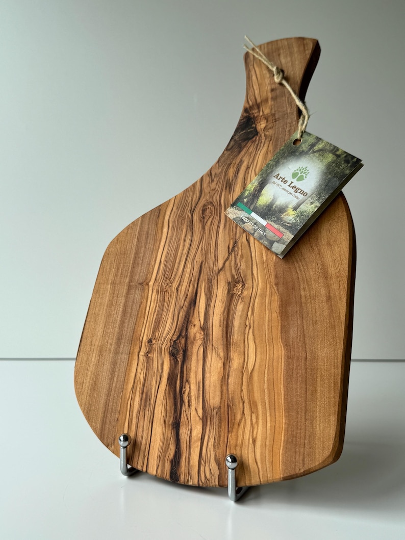 May include: A wooden cutting board with a handle, featuring a natural wood grain pattern. The board has a tag that reads "Arte Legno" and "Product of Italy". The board is supported by two metal stands.