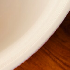 May include: Close-up of the bottom edge of a white ceramic bowl. The bowl has a smooth, rounded edge and a slight lip.