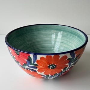 Puede incluir: Un cuenco de cerámica con interior turquesa y diseño floral. El exterior presenta grandes flores naranjas con centros negros, hojas azules y un borde azul oscuro. El cuenco se utiliza probablemente para servir o exhibir.