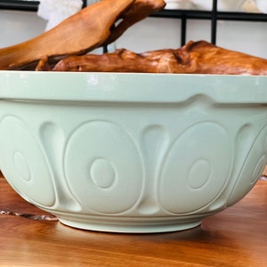 May include: A large, light green ceramic mixing bowl with a fluted design. The bowl is sitting on a wooden surface.