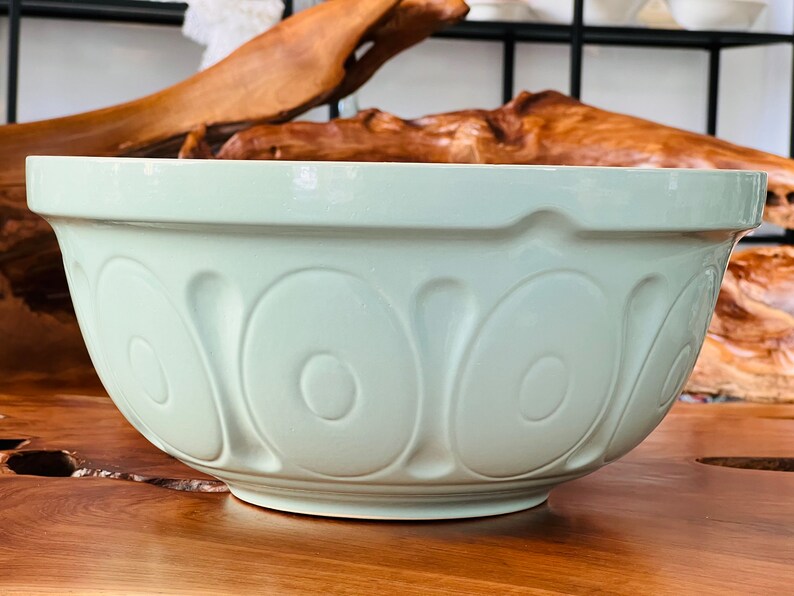 May include: A large, light green ceramic mixing bowl with a fluted design. The bowl is sitting on a wooden surface.