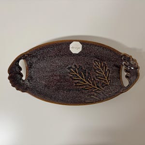 May include: Oval-shaped, dark brown ceramic serving tray with a speckled surface and leaf design. The tray has integrated handles and a small white sticker with text. A decorative piece for serving.