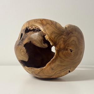 Könnte beinhalten: Eine handgeschnitzte Holzschale mit einer einzigartigen, organischen Form. Die Schale besteht aus hellbraunem Holz mit sichtbarer Maserung und natürlichen Löchern. Der Innenraum ist hohl und bildet ein skulpturales Objekt.