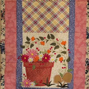 May include: A quilted table runner featuring a floral design. The center panel displays a flower pot overflowing with embroidered flowers in shades of pink, orange, and white. The runner is bordered with pink and blue patterned fabric.