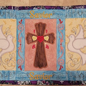 May include: A blue and yellow quilted table runner with embroidered doves, lilies, and a cross. The text "Savior" and "He is Risen" is embroidered on the runner.
