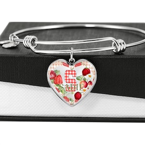 May include: A silver bangle bracelet with a heart-shaped charm. The charm displays a strawberry design and the name "SADIE" in green. The background has a red and white gingham pattern.