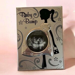 May include: A rectangular silver-toned picture frame featuring a black silhouette of a person with a ponytail. The frame includes the text Baby Bump and a circular cutout displaying a sonogram image, decorated with small flower and swirl patterns.