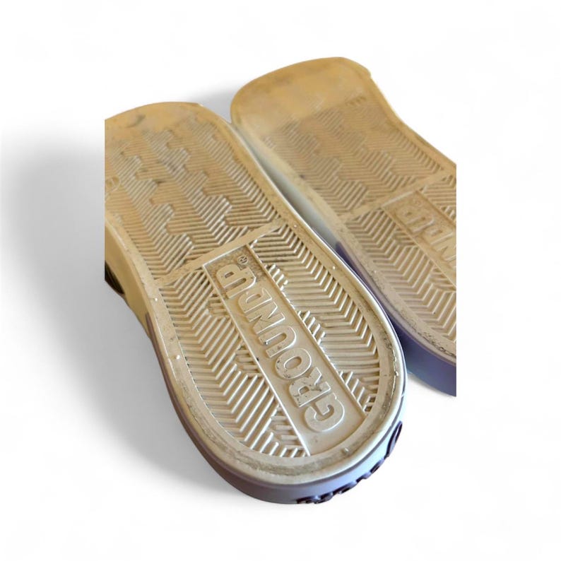 May include: Close-up of the soles of two sneakers. The soles are white with a textured pattern and the word "GROUND UP" is embossed on one sole. The edges of the shoes are purple.