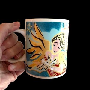 May include: A white ceramic mug featuring a colourful design of a woman holding a dove. The design has a blue background with gold and red accents.