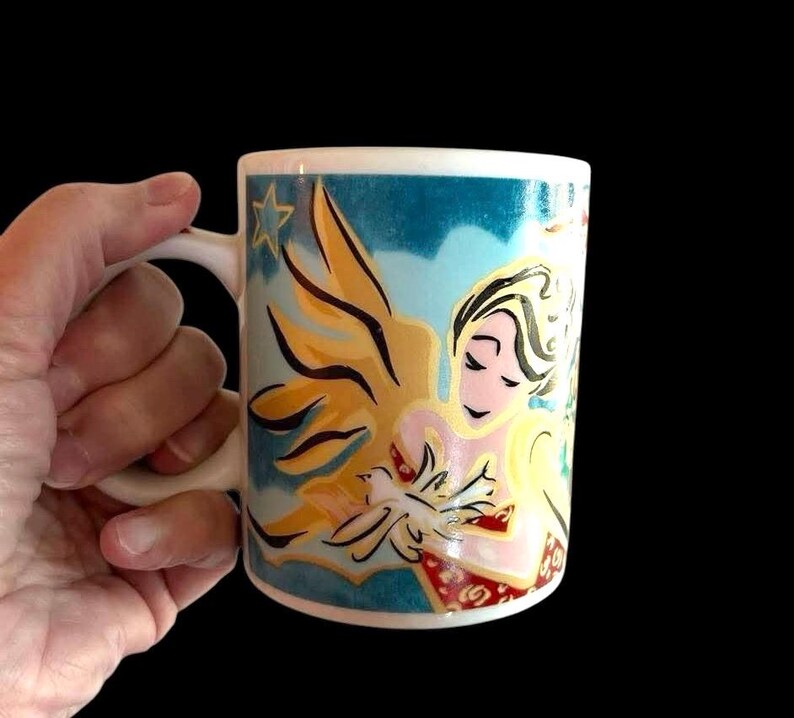 May include: A white ceramic mug featuring a colourful design of a woman holding a dove. The design has a blue background with gold and red accents.