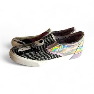 May include: Pair of slip-on sneakers featuring a black and white striped design with a Jack Skellington graphic. The shoes have a white sole, iridescent accents, and a light purple heel.