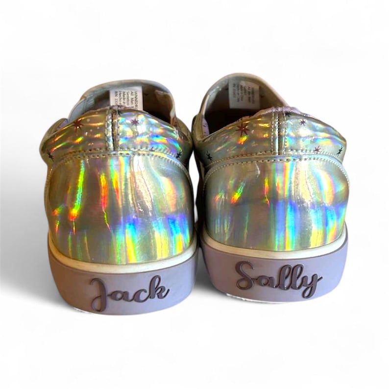 May include: A pair of iridescent silver slip-on sneakers. The shoes have a holographic sheen and are decorated with small star-shaped accents. The word "Jack" is written on the heel of one shoe, and "Sally" on the other.