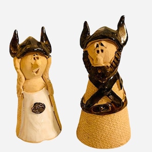 May include: Two ceramic Viking figurines. One is a woman in a white dress, the other a man with a beard. Both wear horned helmets and have textured, tan-colored bodies. The woman has her hands up near her face.