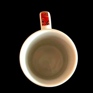 May include: White ceramic coffee mug with a red, orange and yellow patterned handle.