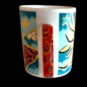 May include: White ceramic mug with a colourful design featuring a sun, stars, and clouds. The handle is red with a swirling pattern.