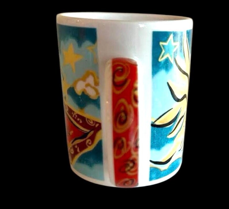 May include: White ceramic mug with a colourful design featuring a sun, stars, and clouds. The handle is red with a swirling pattern.