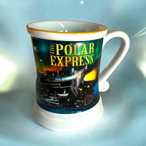 May include: A white ceramic mug featuring a graphic of a steam locomotive and the text THE POLAR EXPRESS in yellow lettering. The mug has a wide handle, a flared rim with a thin yellow border, and a solid white base, showcasing a dark scene.