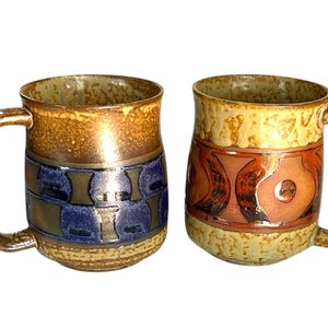 May include: Two ceramic mugs with handles. One mug features a blue and brown geometric design, while the other has a red and orange pattern. Both mugs have a textured, glazed finish and a brown handle.