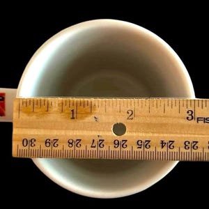 May include: A white ceramic coffee mug with a red and orange floral design on the handle. The mug is sitting on a wooden ruler with the numbers 1, 2, and 3 visible. The ruler is labelled "Fiskars".