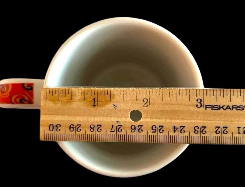 May include: A white ceramic coffee mug with a red and orange floral design on the handle. The mug is sitting on a wooden ruler with the numbers 1, 2, and 3 visible. The ruler is labelled "Fiskars".