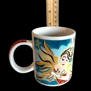 May include: White ceramic mug featuring a blue, gold, and red angel design. The angel is holding a white dove. The mug has a star in the top left corner of the design.