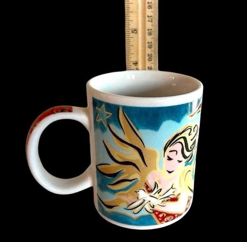 May include: White ceramic mug featuring a blue, gold, and red angel design. The angel is holding a white dove. The mug has a star in the top left corner of the design.