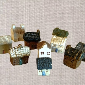 May include: A collection of miniature ceramic houses in various colors and styles. The houses feature details like chimneys, thatched roofs, and painted doors. The color palette includes beige, brown, green, and white.