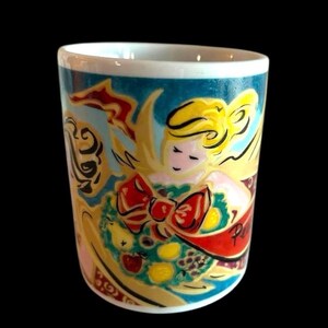 May include: A white ceramic mug featuring a colourful illustration of an angel holding a wreath of fruit and a red ribbon. The angel has blonde hair and a pink face. The background is a teal blue colour.