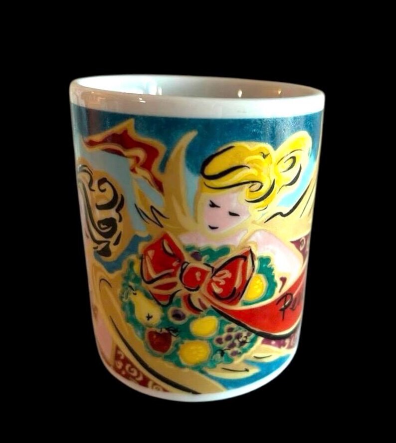 May include: A white ceramic mug featuring a colourful illustration of an angel holding a wreath of fruit and a red ribbon. The angel has blonde hair and a pink face. The background is a teal blue colour.