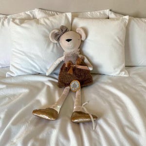 May include: A plush mouse doll with a beige body, a brown dress, and gold sequined feet. The doll has a bow on its head and a small decorative emblem on its leg. The doll is sitting on a white bed with white pillows.
