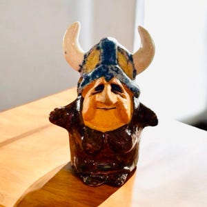 May include: A ceramic figurine of a Viking with a horned helmet. The figure has a tan face, a blue and yellow helmet, and brown clothing. The horns are a cream color. The figurine is on a wooden surface.