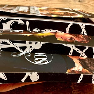 May include: A stack of four paperback books with black covers and white text. The books feature a nautical theme with anchors, chains, and the word "SUS" on the bottom book. The top book has the title "The Mermaid's Revenge" and the bottom book has the title "The Siren's Song, Book 1".
