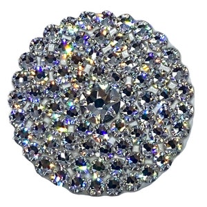 May include: A round, silver-toned, iridescent rhinestone embellishment with a circular shape. The embellishment is made up of many small, round rhinestones that are closely packed together.
