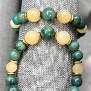 May include: Two bracelets with green, yellow, and brown beads. The bracelets are stacked on a gray fabric surface. The top bracelet has a gold clasp. The bottom bracelet has a clear crystal bead in the center.