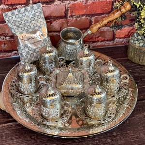May include: A silver Turkish coffee set comprising six cups, saucers, and a sugar bowl on a decorative tray. Includes a coffee pot and a bag of "Kocatepe Türk Kahvesi" coffee. The set is set against a brick background.