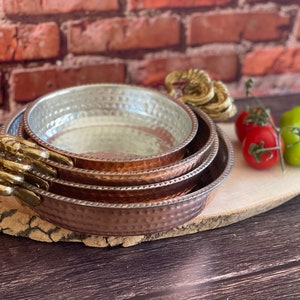 Può includere: Un set di quattro piatti da portata in rame martellato con manici color oro. I piatti sono disposti su un tagliere di legno, con pomodori freschi e un peperone verde. Il rame ha una calda tonalità marrone-rossastra.