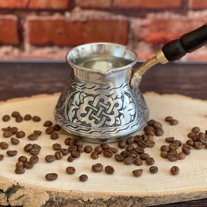 May include: A silver coffee pot with an ornate design and a dark wooden handle, resting on a wooden slab. Coffee beans are scattered around the pot. The pot has a brass handle attachment.