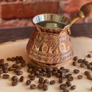May include: A copper Turkish coffee pot with intricate embossed patterns and a wooden handle, filled with liquid. Coffee beans are scattered around the pot on a wooden surface, suggesting a coffee preparation theme.