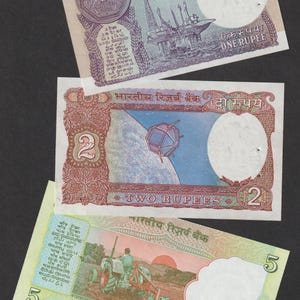 Puede incluir: Se muestran tres billetes indios sobre un fondo negro. Los billetes son de 1, 2 y 5 rupias. El billete de 1 rupia presenta una plataforma petrolífera, el de 2 rupias un satélite y el de 5 rupias un tractor.