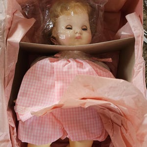 May include: A vintage baby doll in a box, wrapped in pink tissue paper. The doll has blonde hair, closed eyes, and a pink gingham dress. The doll's shoes are pink with small bows.