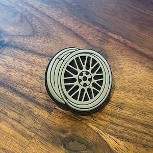Puede incluir: Un pin de metal de color plateado con forma de rueda de coche con un neumático negro. La rueda tiene un diseño detallado con radios y un tapacubos central.