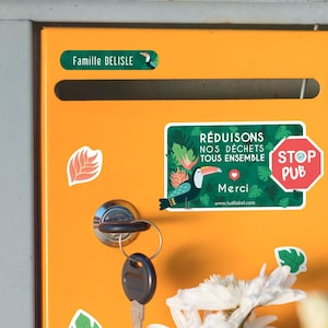 Könnte beinhalten: Ein gelber Briefkasten mit einem Schild, auf dem "Reduisons nos déchets tous ensemble" und "Stop Pub" steht. Der Briefkasten hat ein Schlüsselloch und einen Aufkleber mit der Aufschrift "Famille DeLisle".