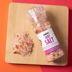 Peut inclure: Un pot en verre de sel rose de l'Himalaya avec des flocons de piment rouge, étiqueté "Ember's Hot Salt". Le pot a un moulin sur le dessus et est posé sur une planche à découper en bois.