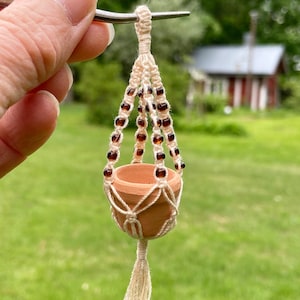 Może przedstawiać: Mały gliniany doniczek wiszący na makramowym haczyku do roślin z brązowymi koralikami. Haczyk ma biały frędzl.
