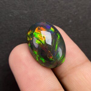 Op de afbeelding: Ovaalvormige zwarte opaal edelsteen met levendig intern vuur. De opaal toont flitsen van groen, oranje en blauw, waardoor een dynamisch kleurenspel ontstaat. Het gepolijste oppervlak reflecteert licht.