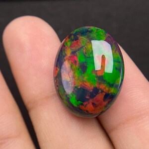 Puede incluir: Una piedra preciosa de ópalo pulida, de forma ovalada. El ópalo muestra un vibrante juego de colores, con destellos de verde, rojo y naranja sobre una base oscura. La gema se sostiene entre los dedos, mostrando su superficie reflectante.