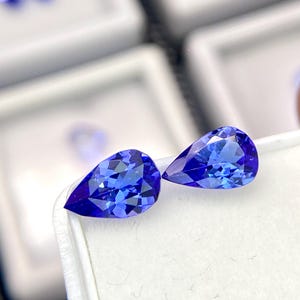 May include: Two pear-shaped blue gemstones, also known as teardrop-shaped, are displayed in a white box. The gemstones are a vibrant blue color and have a smooth, polished surface.