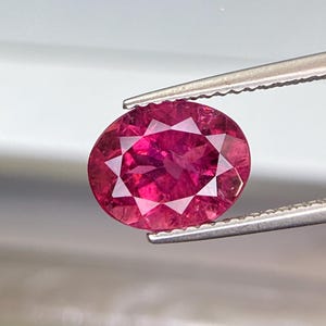 Puede incluir: Una gema facetada, de forma ovalada, de color rojo intenso, es sostenida por unas pinzas. La gema tiene un corte brillante con múltiples facetas que reflejan la luz, creando un efecto brillante. El fondo es de un color suave y neutro.